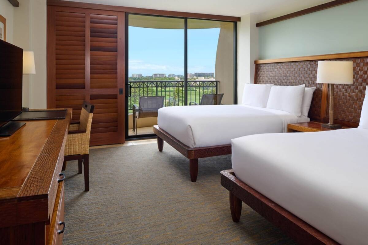 Habitación de hotel con dos camas, un escritorio, una silla, una mesita de noche y vistas a un paisaje verde a través de una puerta corredera.