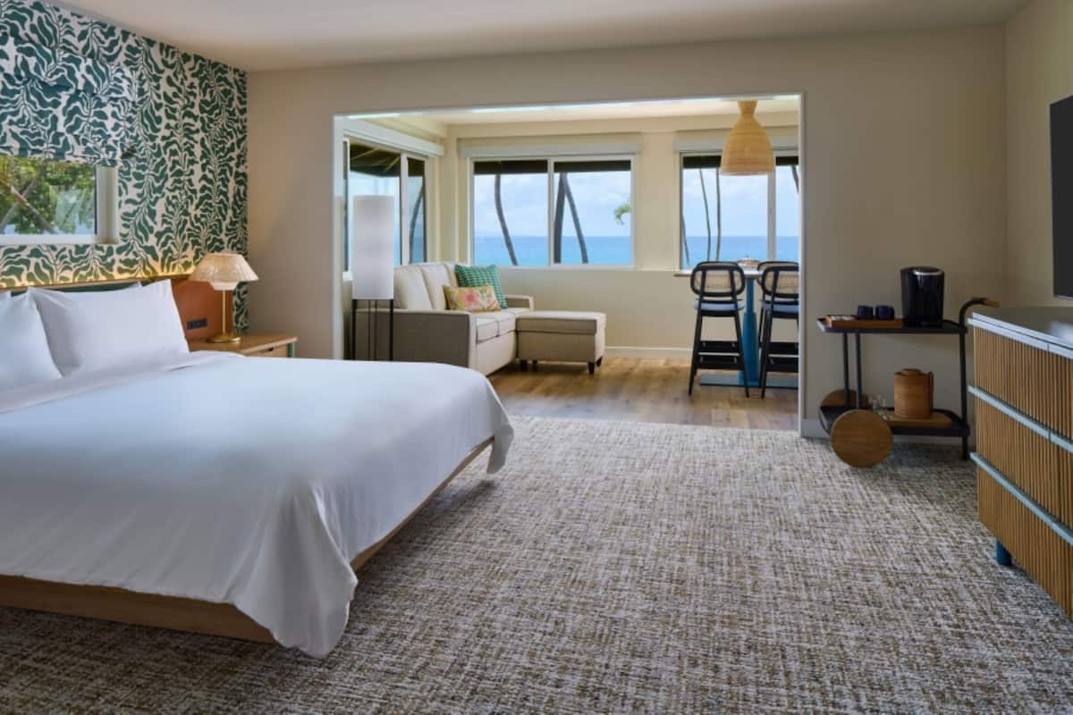Habitación de hotel con una cama grande, un escritorio con silla, vista al océano y un balcón con mesa y sillas.