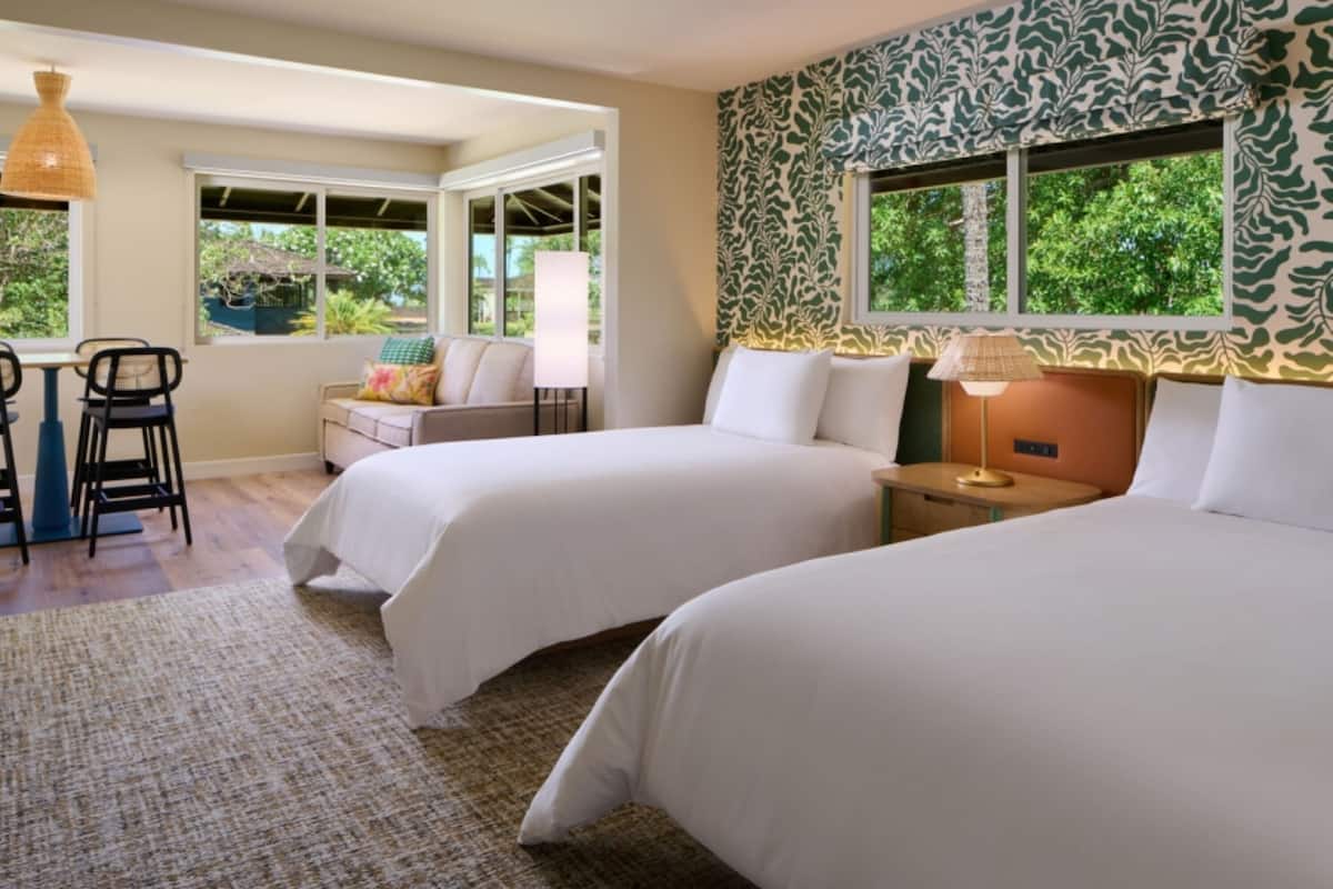 Habitación de hotel con dos camas, un escritorio con sillas, una ventana con vista a la vegetación y una pared con diseño.
