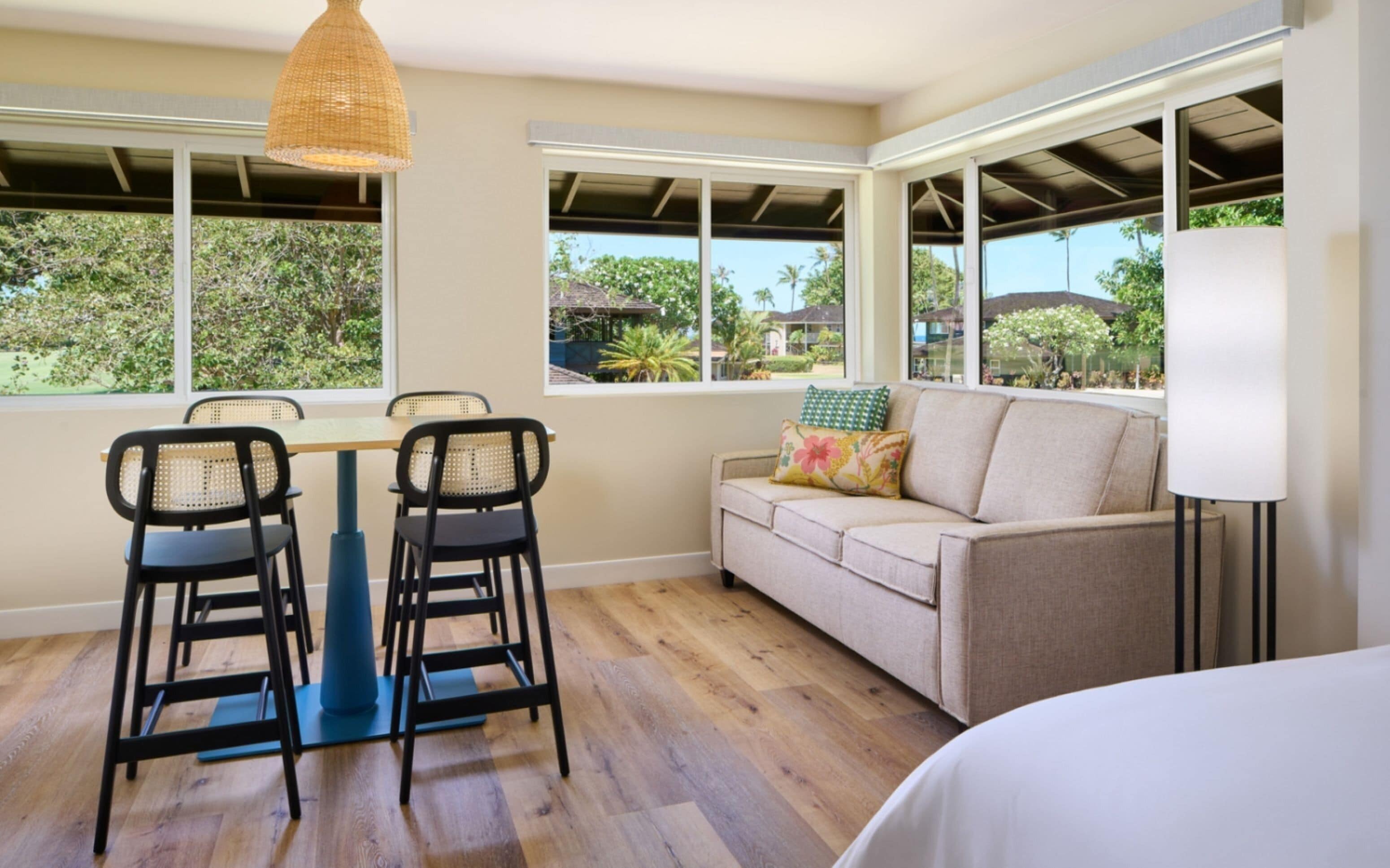 Quarto com piso de madeira, sofá, duas cadeiras, mesa e janelas amplas.