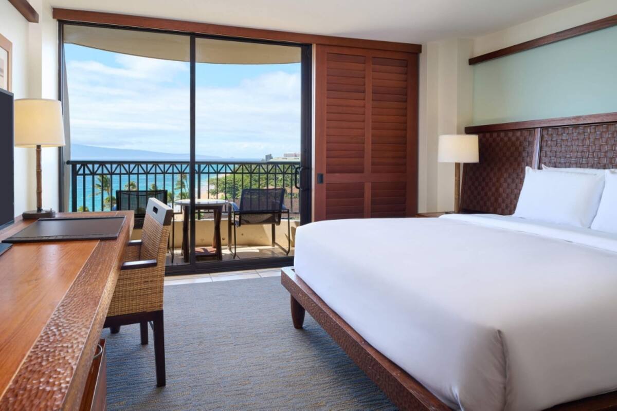 Habitación de hotel con una cama grande, un escritorio y una silla. Tiene un balcón con vistas al océano.