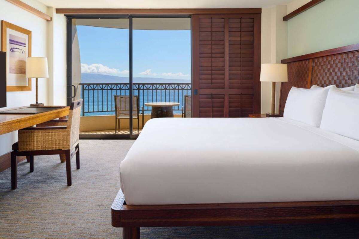 Habitación de hotel con una cama grande, un escritorio con silla, una mesita y vista al océano a través de una puerta corrediza.