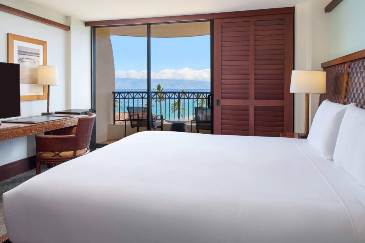 Habitación de hotel con una cama grande, un escritorio con una computadora, una silla y vista al océano a través de una puerta corrediza.