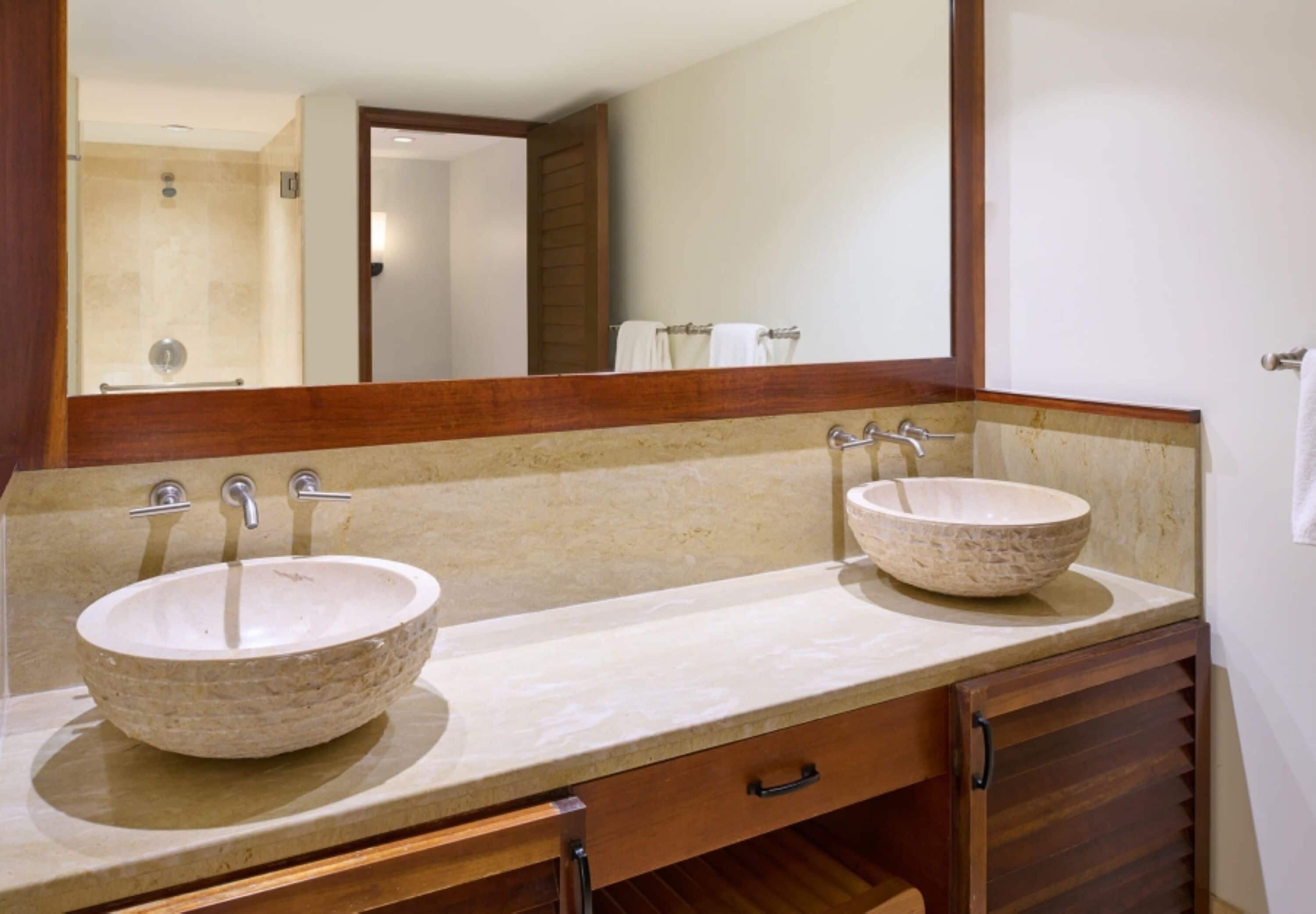 A bathroom with two stone sinks, a wooden cabinet, and a mirror reflecting a shower area.