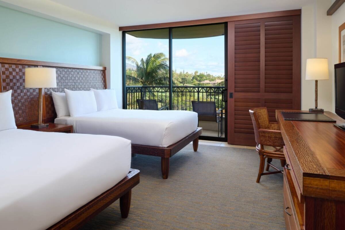 Habitación de hotel con dos camas, un escritorio y un balcón con vistas a palmeras y casas.