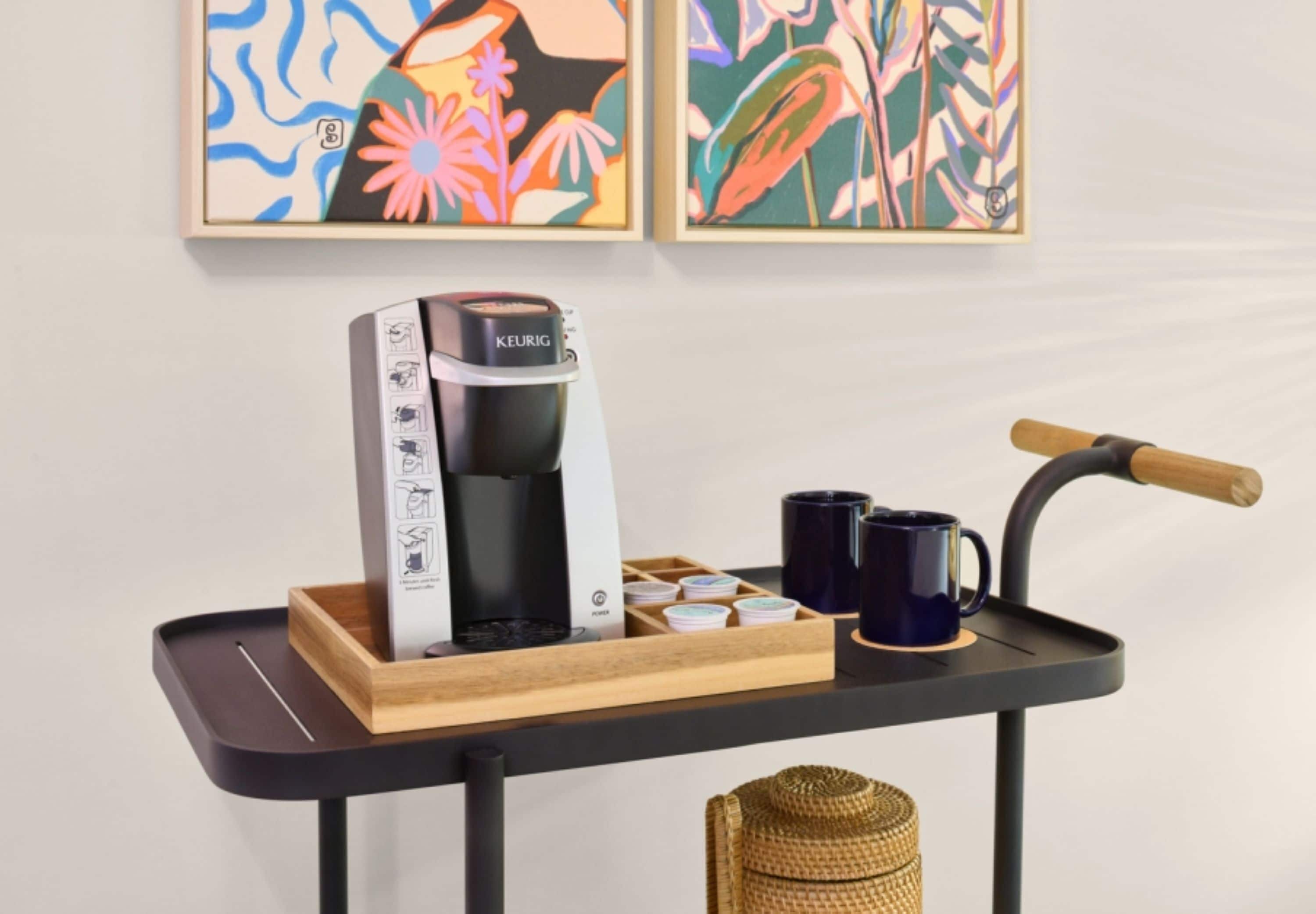Uma cafeteira em um tabuleiro com uma caneca e uma cesta de xícaras.