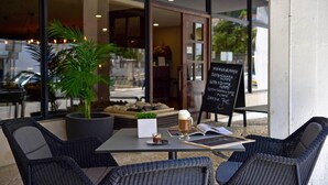Un área de estar al aire libre con sillas de mimbre, una mesa con la carta, una planta en maceta y la entrada a una cafetería.