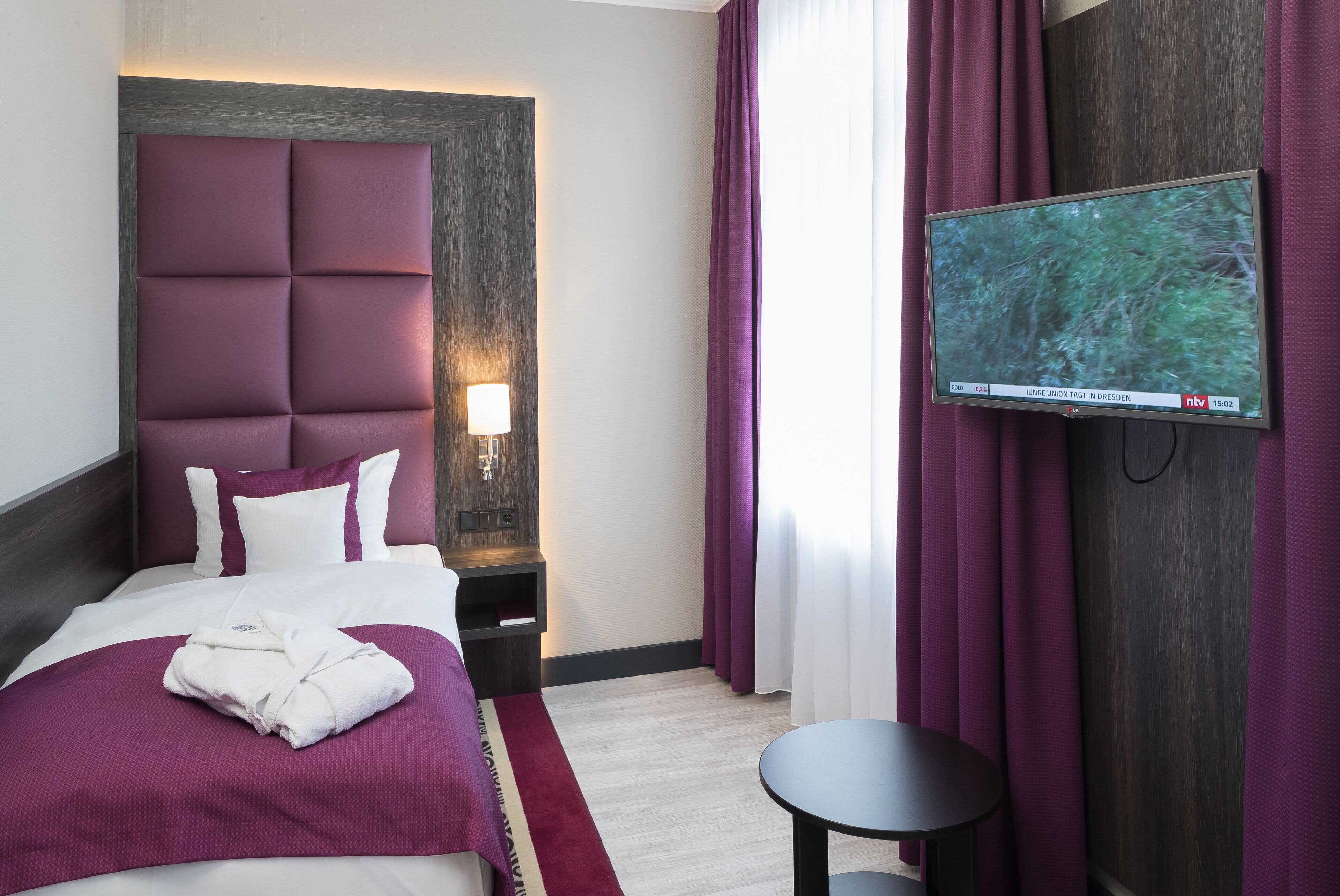 Una habitación de hotel con una cama grande, un televisor de pantalla plana en la pared, una mesita de noche y una pequeña mesa redonda.