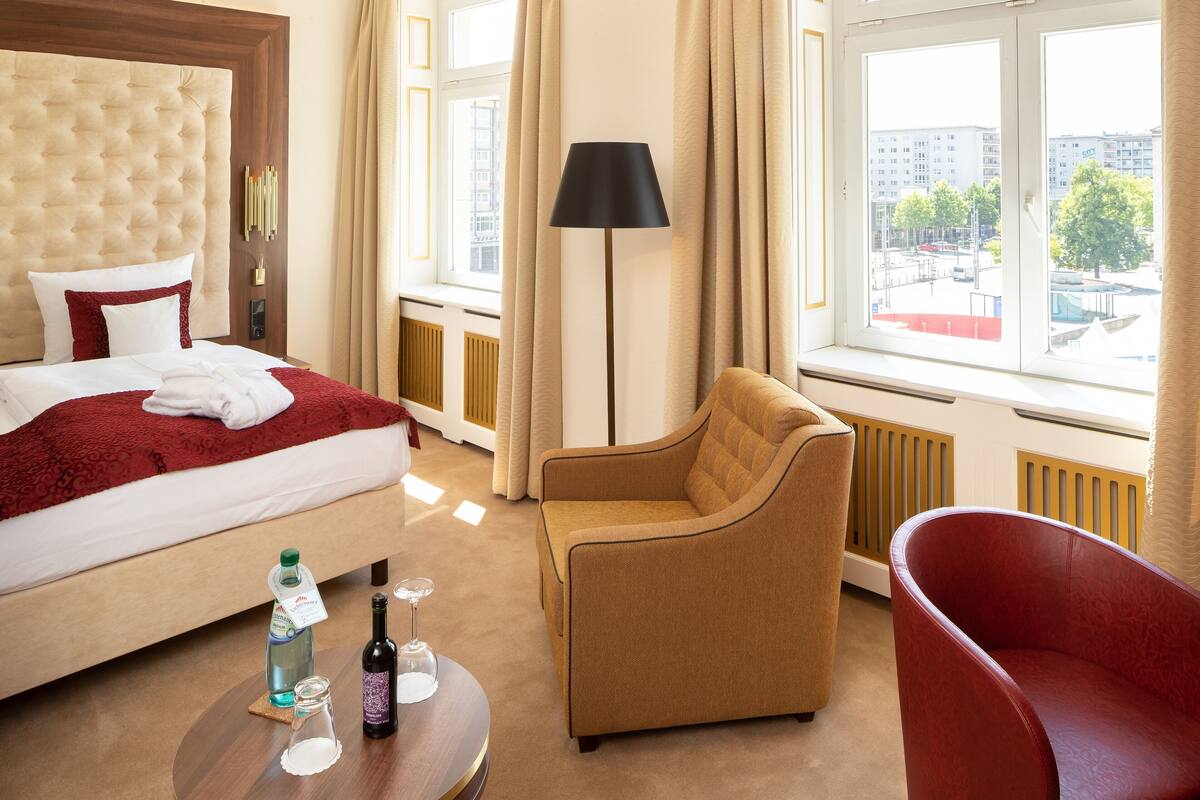 A hotel room with a large bed, a red armchair, a floor lamp, and a view of the city through the window.