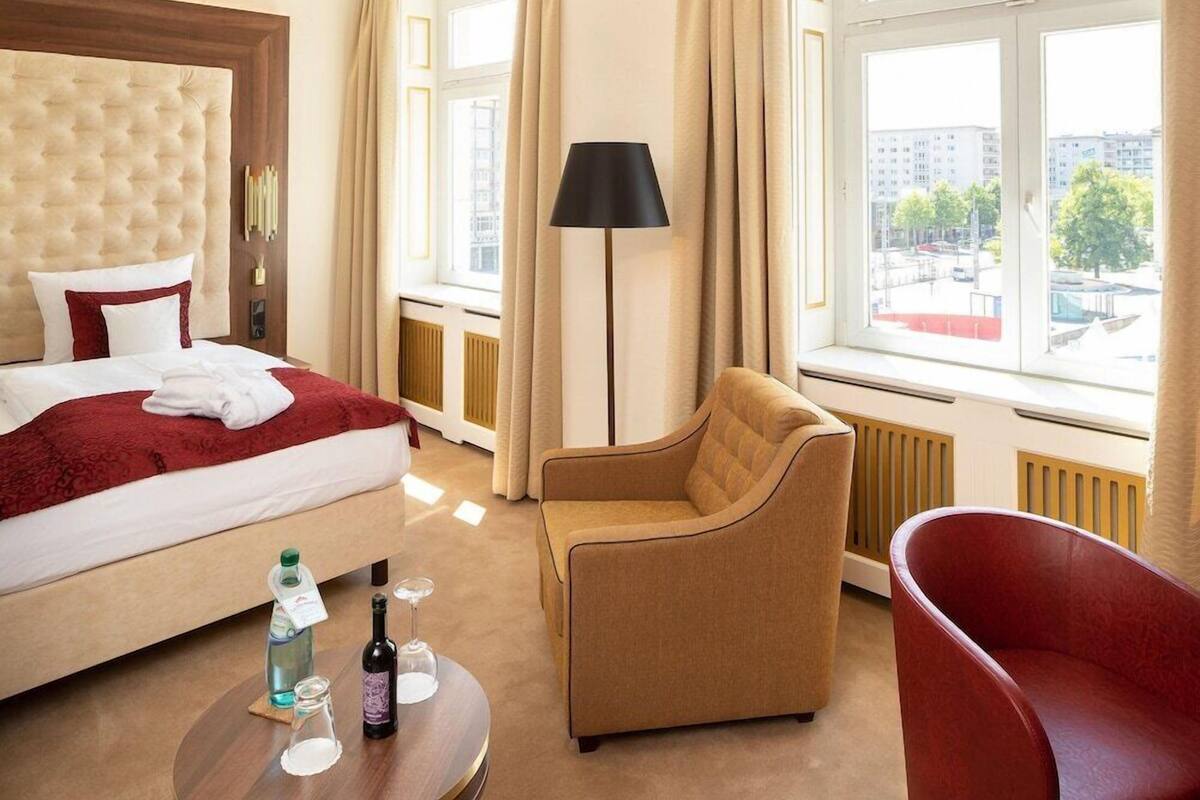 A hotel room with a bed, a red chair, a lamp, and a view of the city.