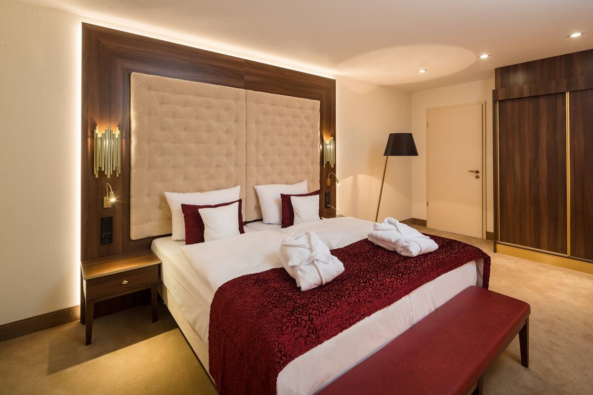 A hotel room with a large bed, a wooden headboard, a bench, a bedside table, and a wardrobe.