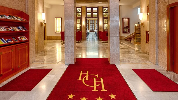 A hotel lobby with a red carpet leading to a grand staircase, marble flooring, and a reception desk with a red chair. - Hotel Chemnitzer Hof (Chemnitz)