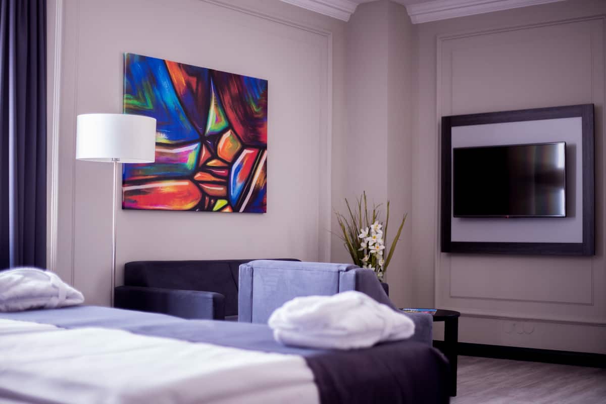 A hotel room with a bed, a TV, a painting, and a vase with flowers.