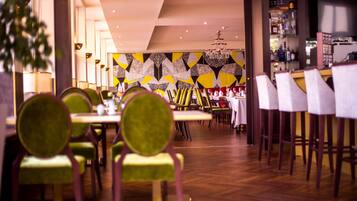 A restaurant interior with a modern design, featuring a geometric patterned wall, wooden flooring, and green upholstered chairs.