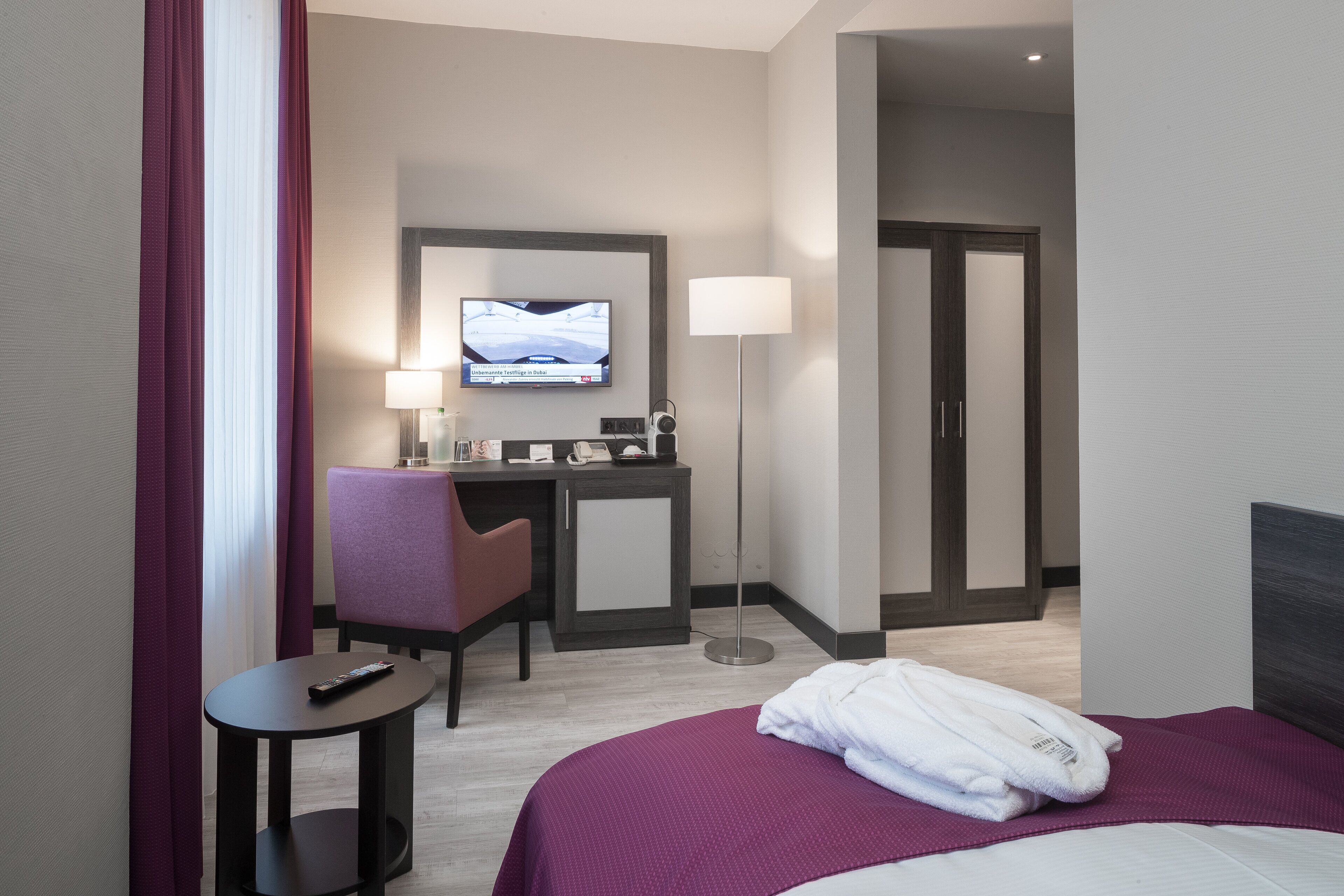 A hotel room with a bed, a desk with a chair, a TV mounted on the wall, and a lamp.