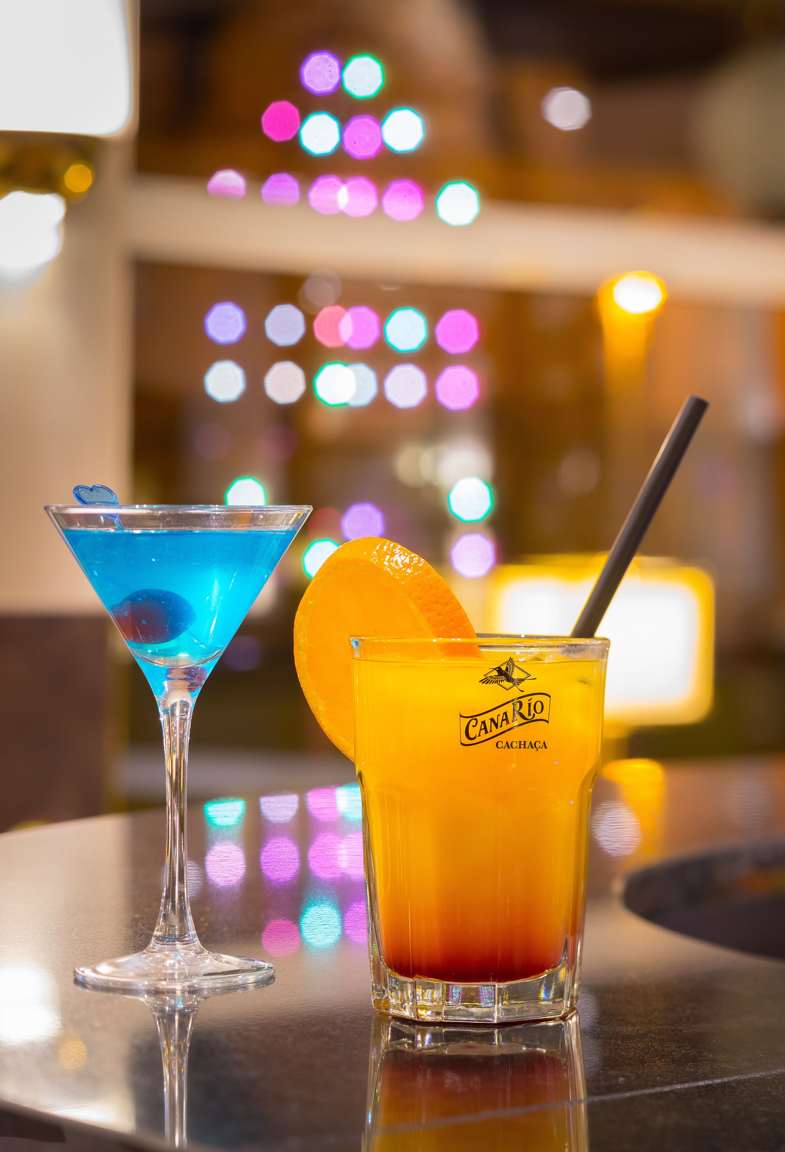 Deux cocktails placés sur une surface réfléchissante, avec un arrière-plan flou évoquant une ambiance festive.