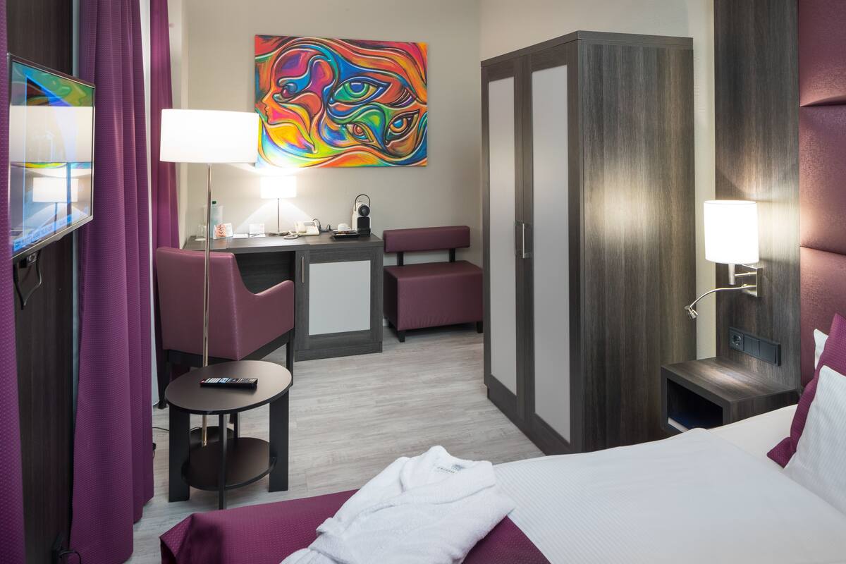 A hotel room with a bed, a desk with a chair, a small table, a robe, and a colorful artwork on the wall.