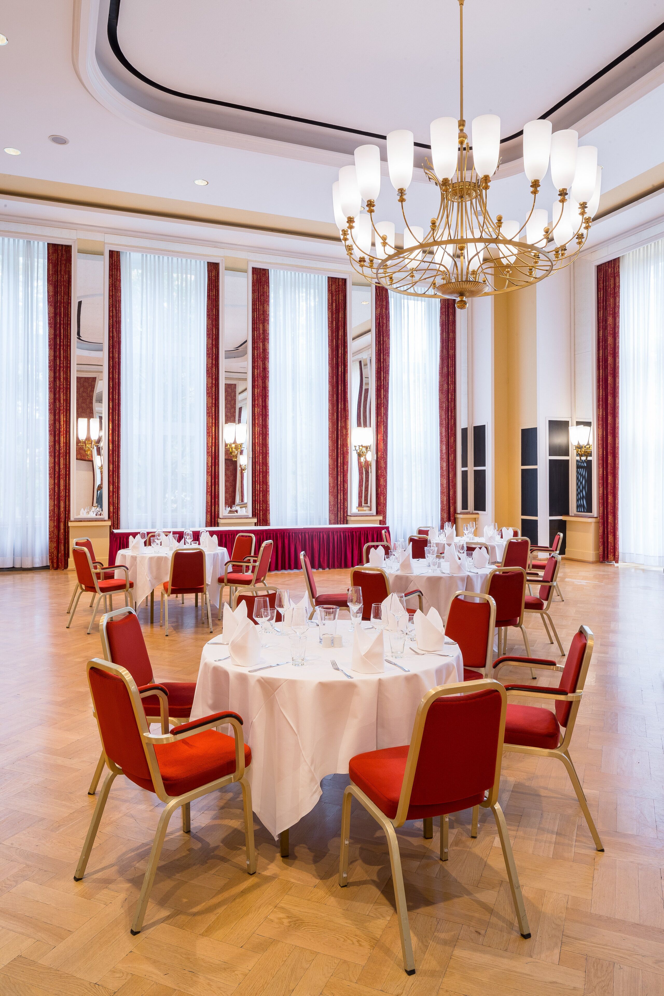 Ein geräumiger Festsaal mit runden Esstischen, roten, gepolsterten Stühlen und einer großen Kristallleuchter.