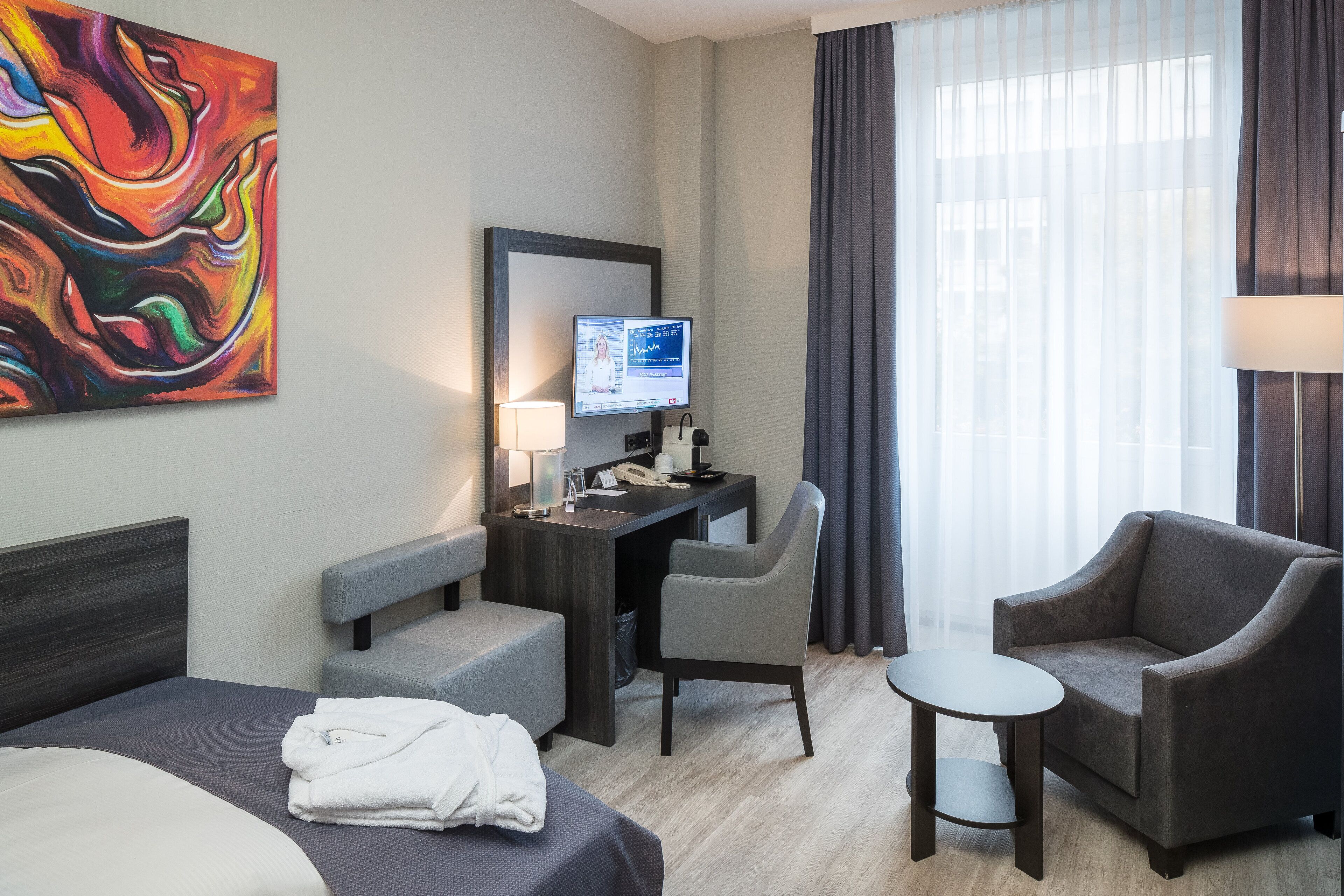 A hotel room with a bed, desk, two chairs, a small table, a TV, and a colorful painting on the wall.