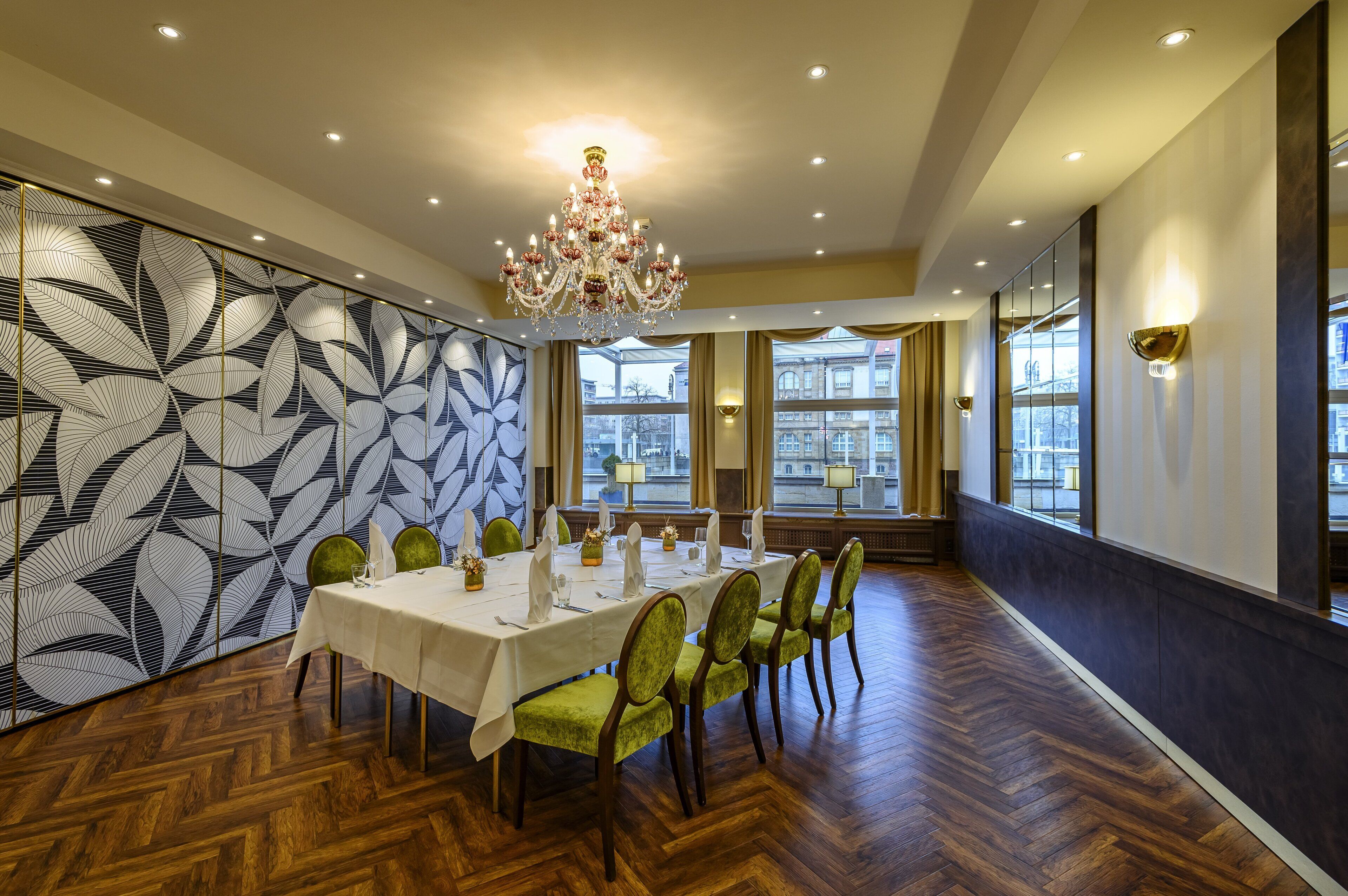 A dining area with a long table, green upholstered chairs, a chandelier, and a patterned wall.