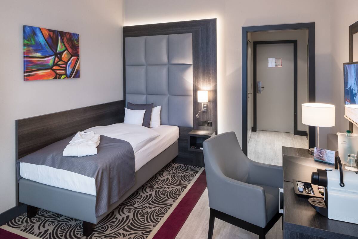 A hotel room with a bed, a chair, a desk, a lamp, a painting, and a door.