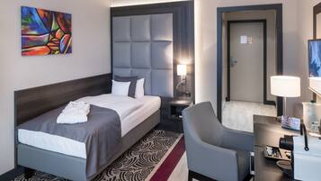 A hotel room with a bed, a chair, a desk, a lamp, a painting, and a door.