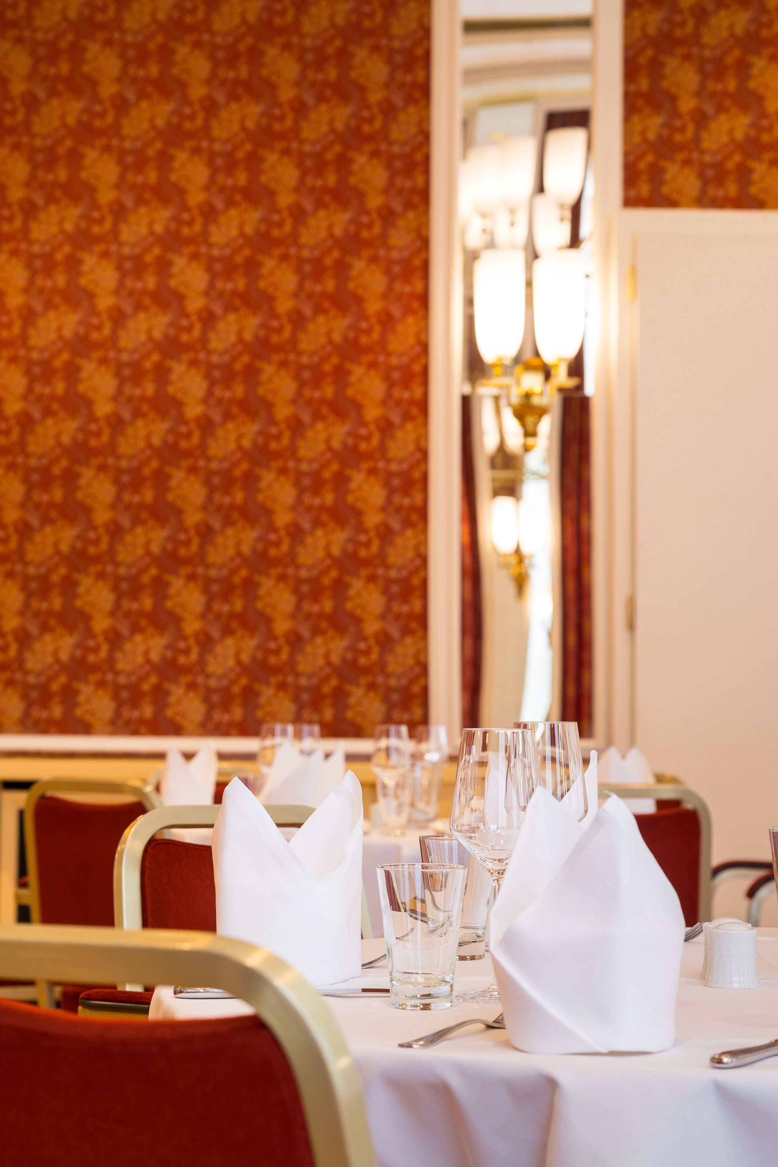 A dining table set with white napkins, glasses, and cutlery, with a patterned wallpaper background and a mirror reflecting a chandelier.