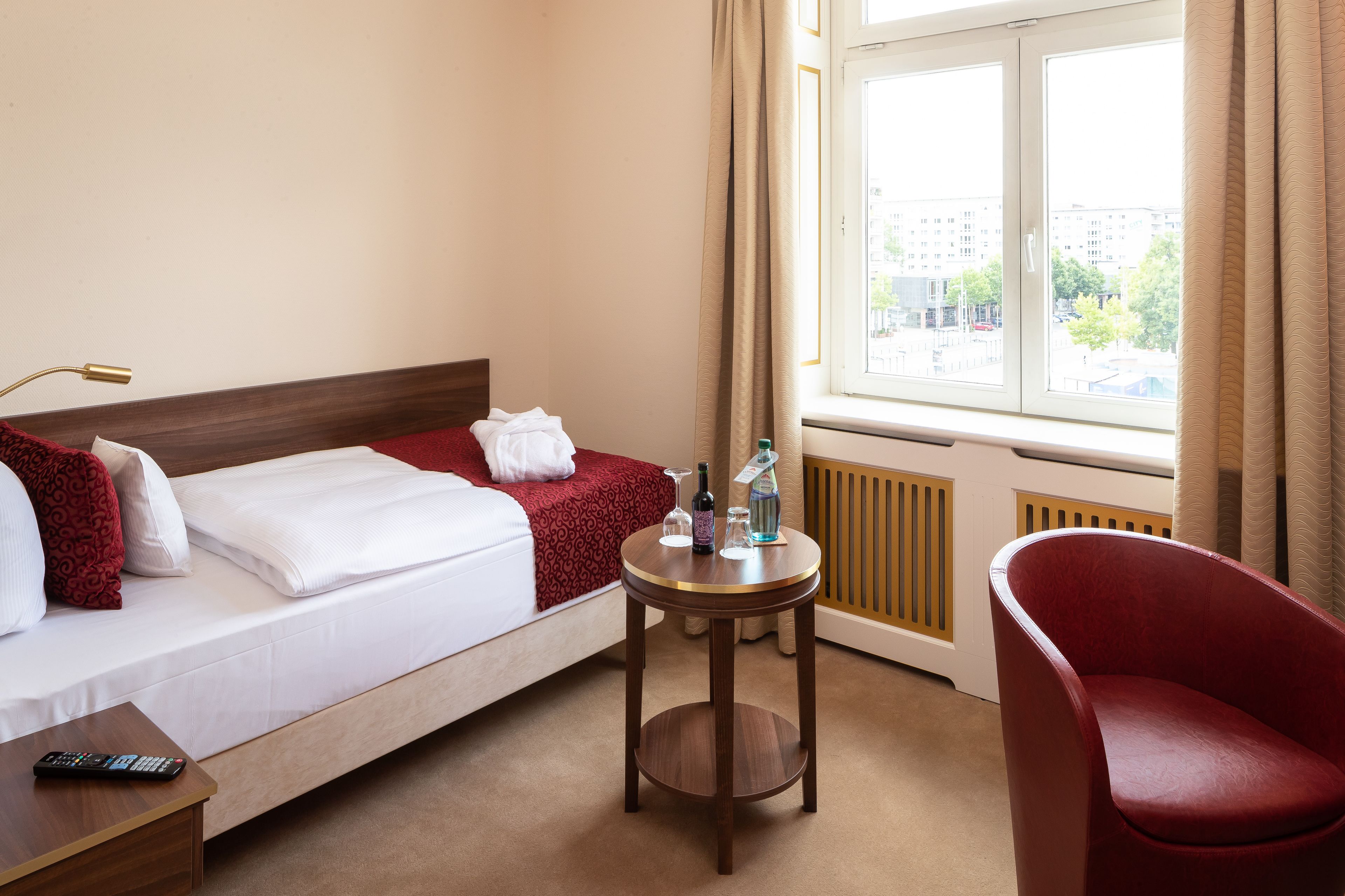 A hotel room with a bed, a small round table with a bottle and glass, a red chair, and a window with a view of buildings.