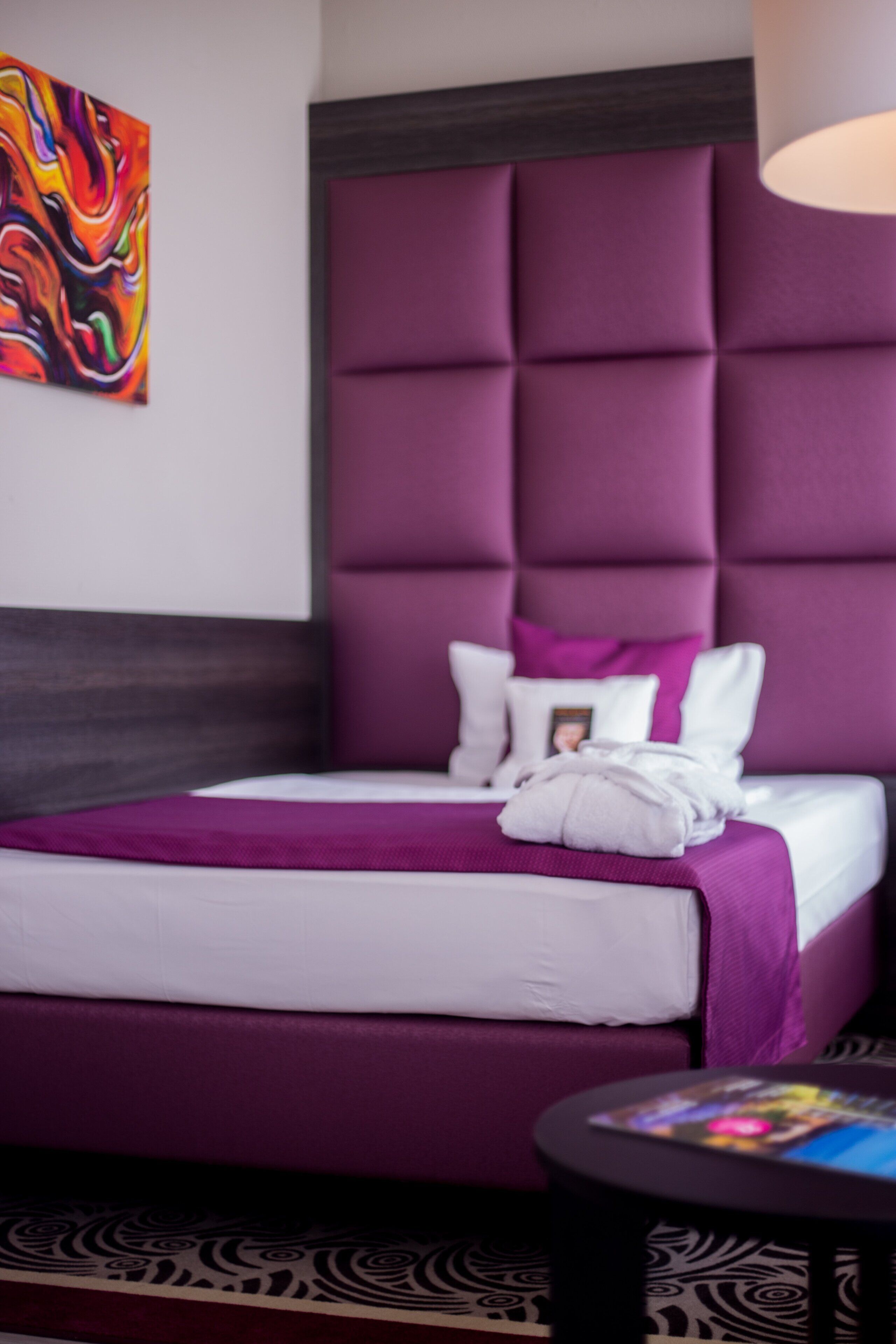 A hotel room with a large tufted headboard, a neatly made bed with a purple bedspread, a small round table, and a colorful abstract painting on the wall.