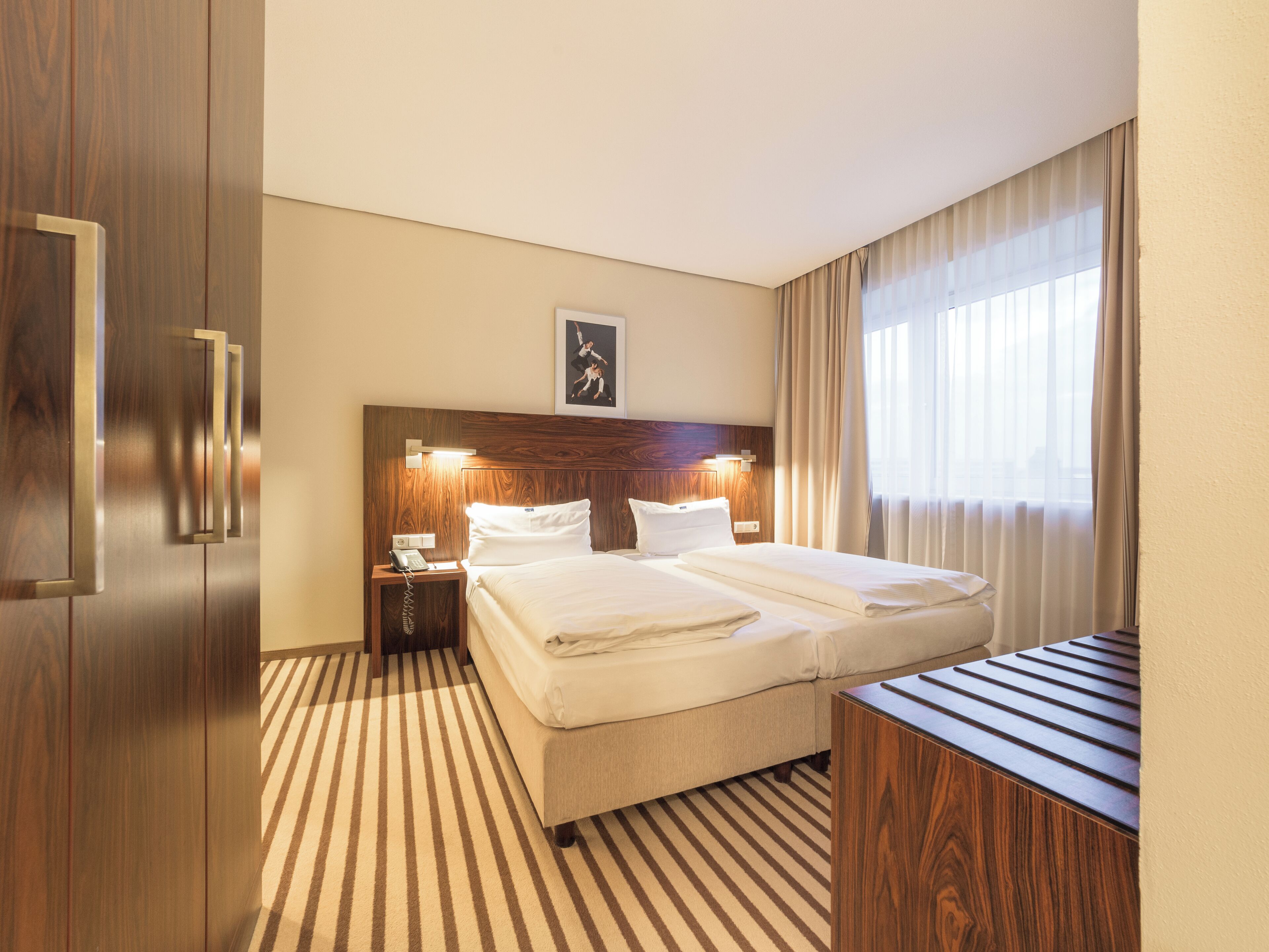 Habitación de hotel con cabecera de madera, cama con ropa de cama blanca, mesita de noche con teléfono y ventana con cortinas.