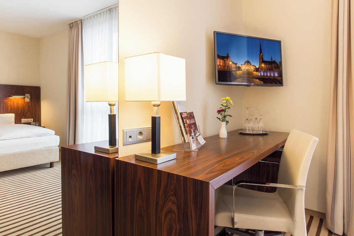 A hotel room with a bed, a desk with a lamp, a chair, a TV on the wall, and a vase with flowers.