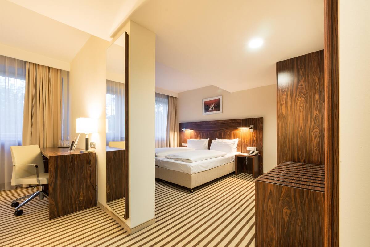 A hotel room with a bed, a desk with a chair, a mirror, and a wooden wardrobe.