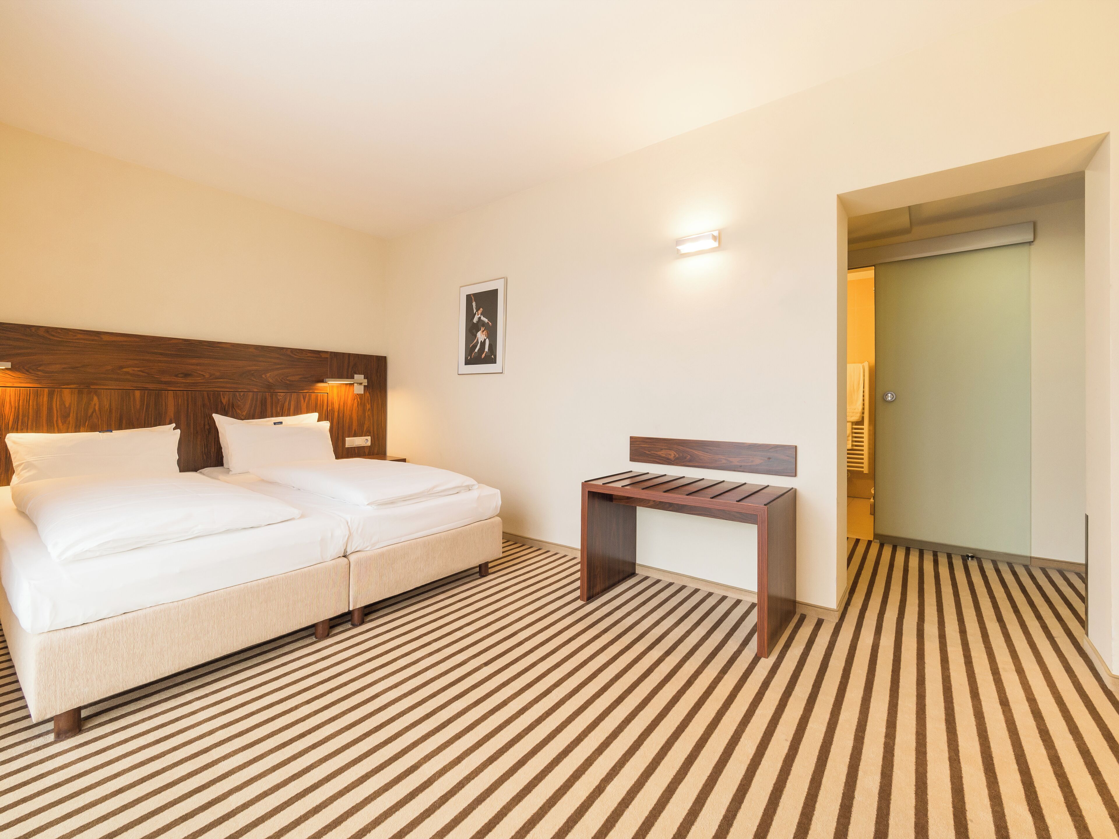 A hotel room with a bed, a small table, a wooden headboard, a wall-mounted light, and a striped carpet.