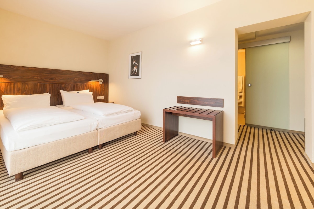 A hotel room with a bed, a small table, a wooden headboard, a wall-mounted light, and a striped carpet.