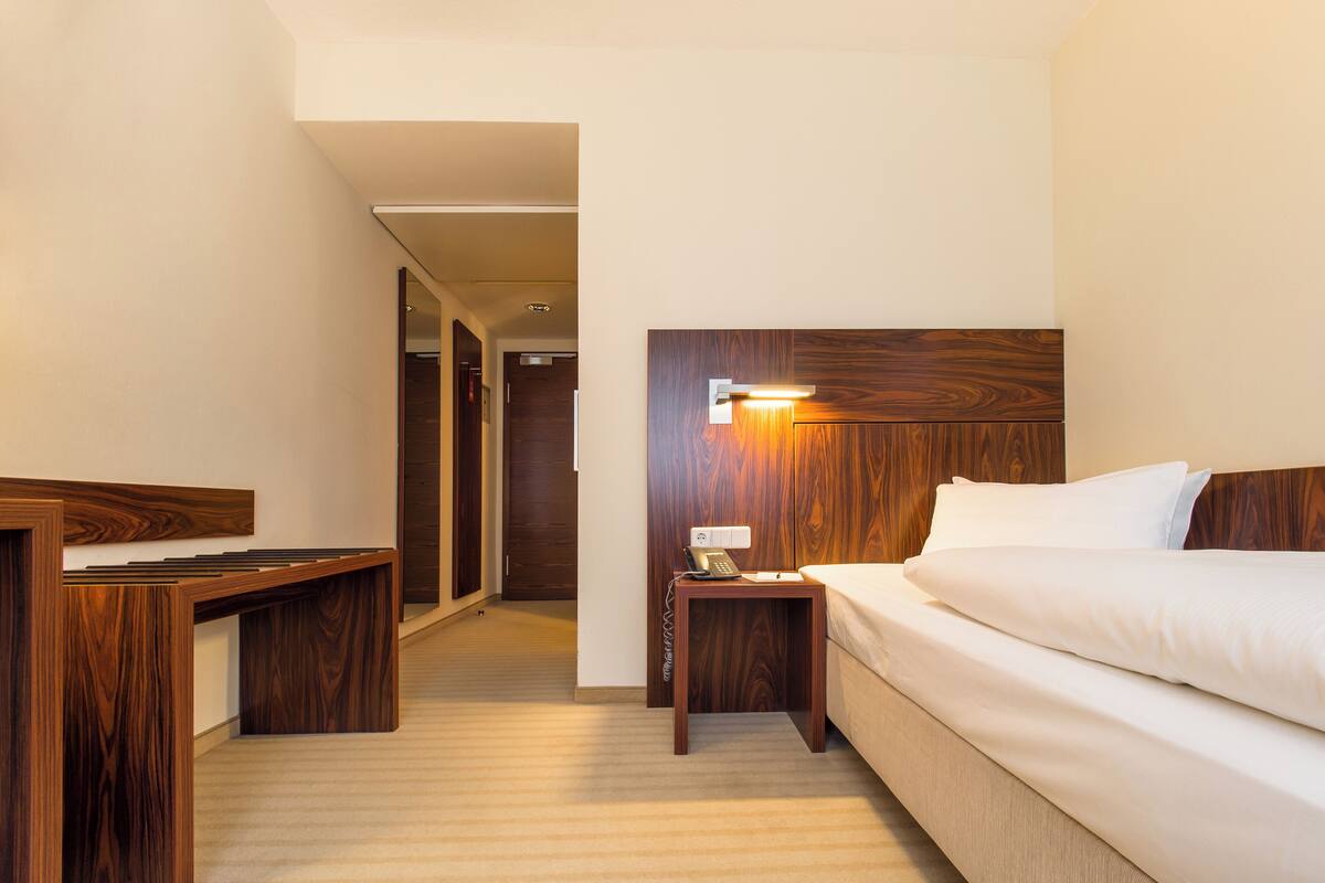 A hotel room with a wooden headboard, a bedside table with a lamp, a desk, and a bed with white bedding.