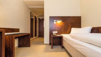 Ein Hotelzimmer mit Holz-Kopfteil, einem Nachttisch mit Lampe, einem Schreibtisch und einem Bett mit weißer Bettwäsche.