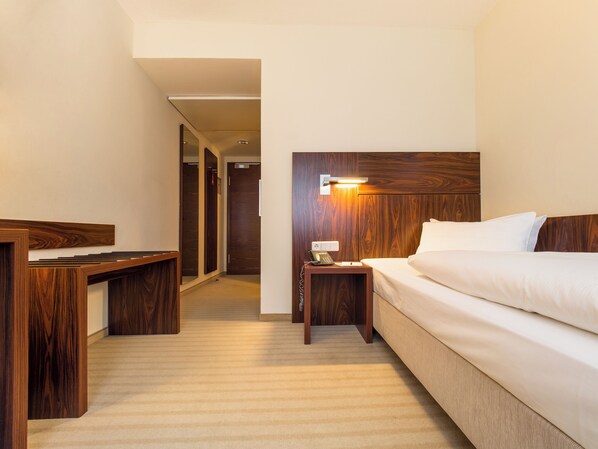 Habitación de hotel con cabecera de madera, mesita de noche con lámpara, escritorio y cama con ropa blanca.