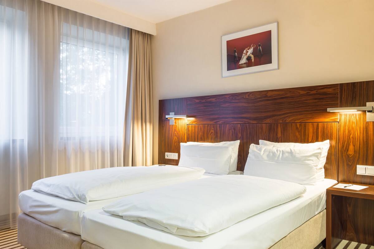 A hotel room with two beds, a wooden headboard, a framed picture on the wall, and a window with sheer curtains.