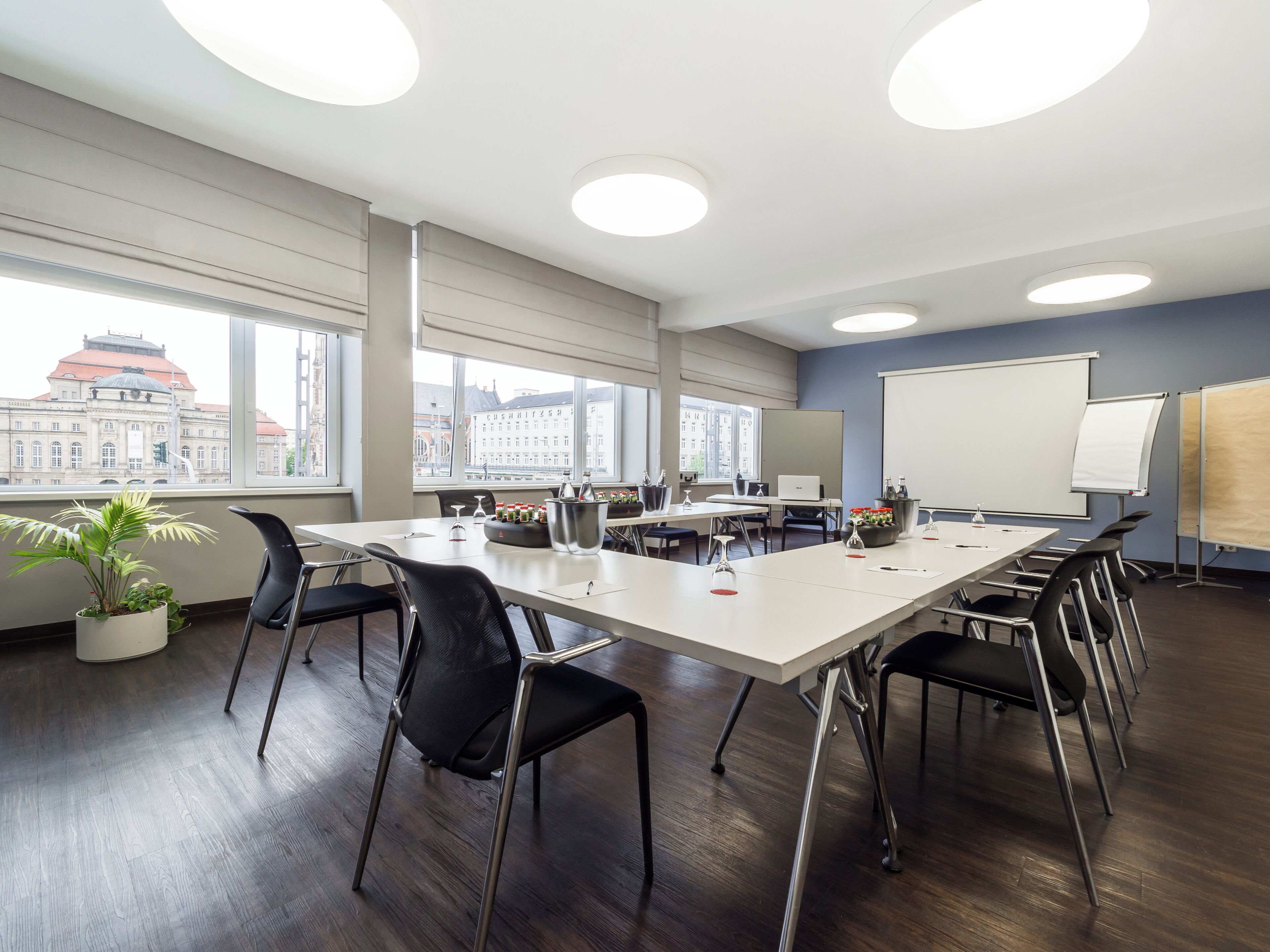 A conference room with a long table, chairs, a projector screen, and a view of buildings through large windows.