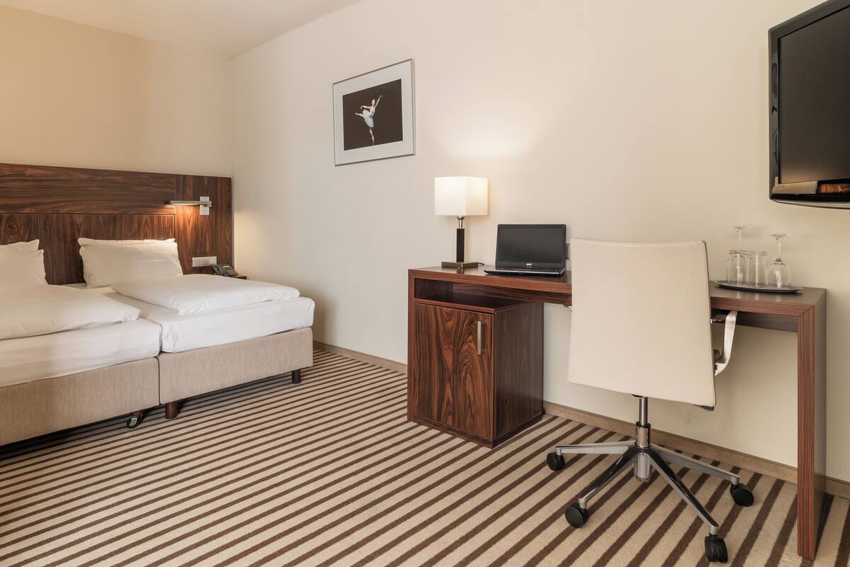 A hotel room with a bed, a desk with a laptop, a chair, a lamp, and a TV on the wall.