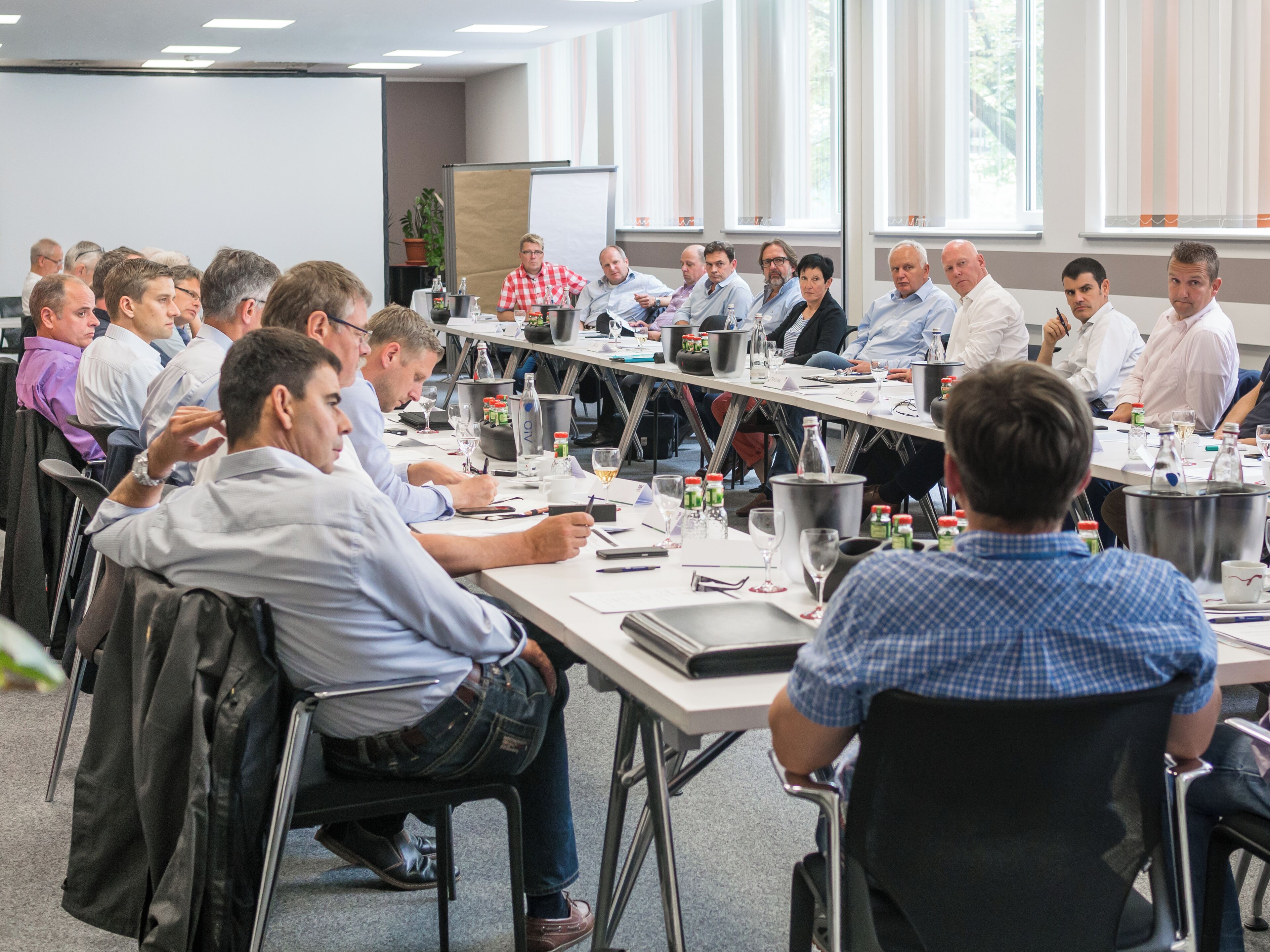 Ein Konferenzraum mit einem langen Tisch, Stühlen und Teilnehmern.