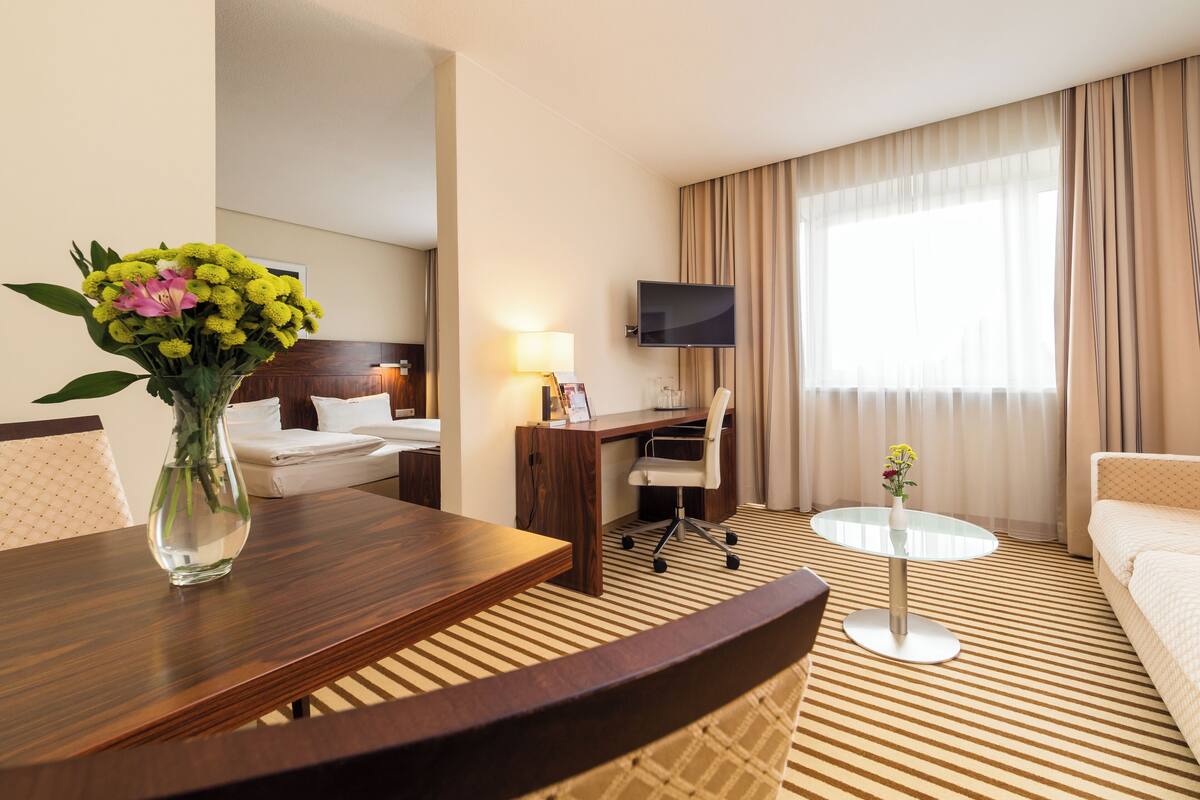 A hotel room with a bed, desk, chair, TV, and a small table with a vase of flowers.