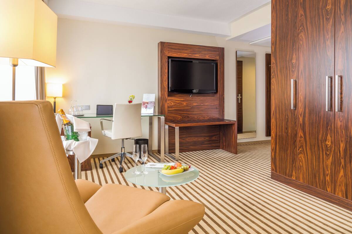 A hotel room with a leather armchair, a glass coffee table with fruit, a flat-screen TV mounted on a wooden panel, a desk with a computer, and a minibar.