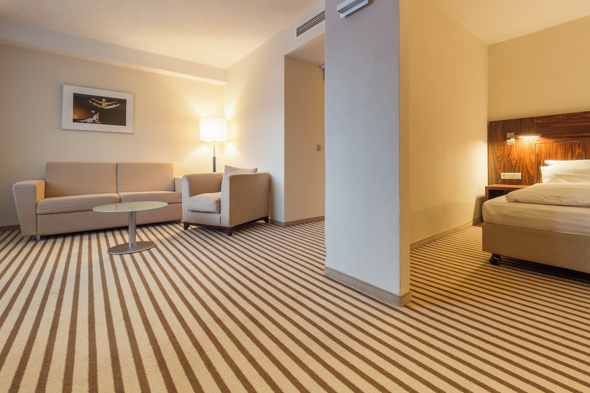 A hotel room with a striped carpet, a sofa, a chair, a small table, a bed, and a nightstand with a lamp.