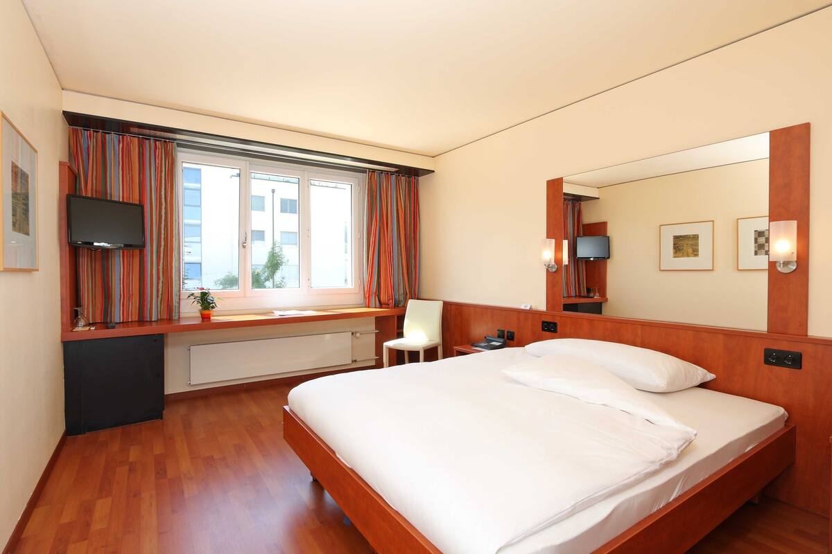 A hotel room with a bed, a TV, a desk with a chair, a window with curtains, and a mirror.