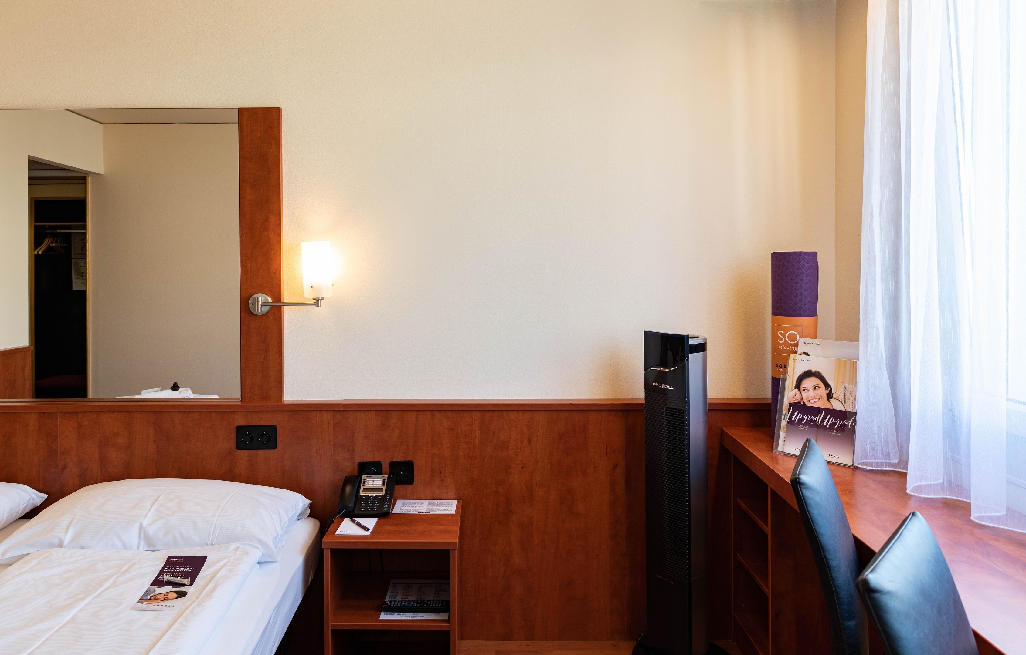 Une chambre d’hôtel avec un lit, un bureau avec un téléphone et des documents, un miroir, une télévision et une fenêtre avec des rideaux.