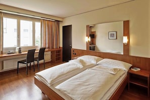 A hotel room with a bed, a desk, two chairs, a window with curtains, a mirror, and a nightstand. - Hotel Arte (Spreitenbach)