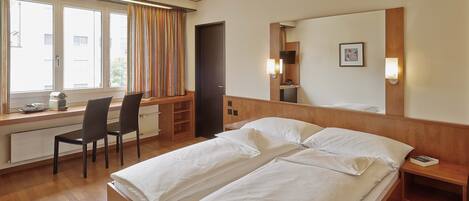 Habitación de hotel con cama doble, escritorio con silla, ventana con cortinas y espejo en la pared.