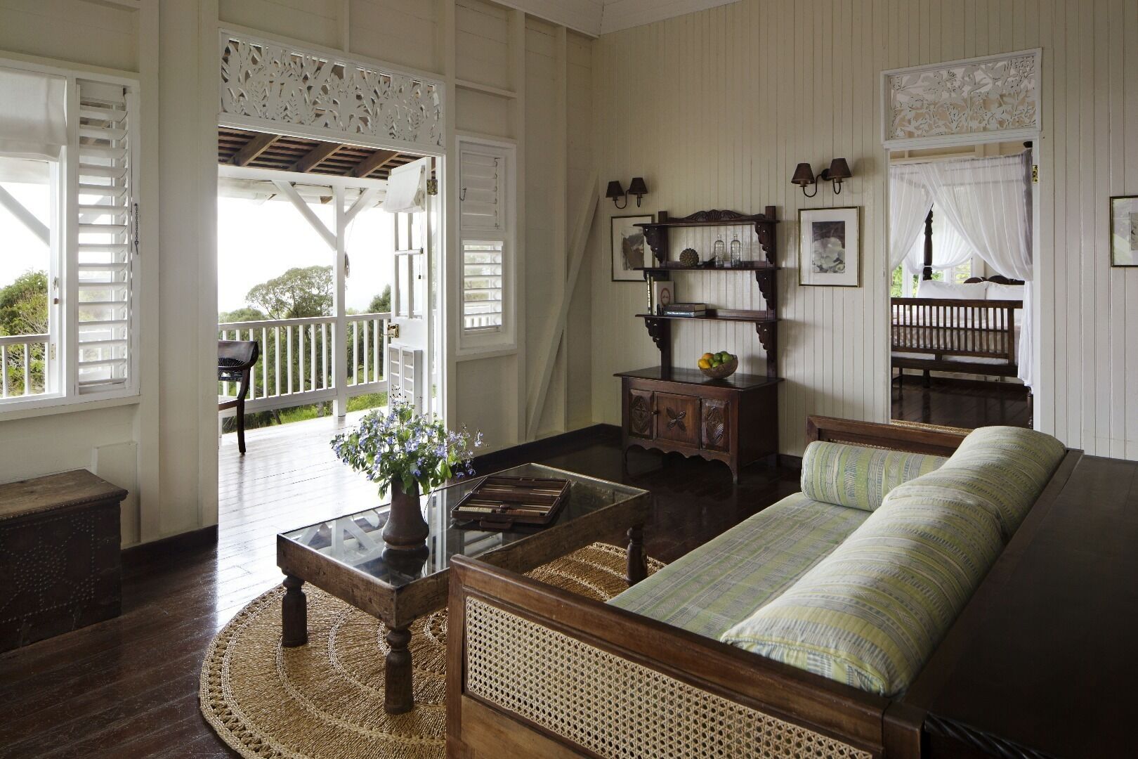 A cozy living room with a sofa, a coffee table with a vase, a wooden cabinet, and a bookshelf. There is a balcony with a railing and a bench.