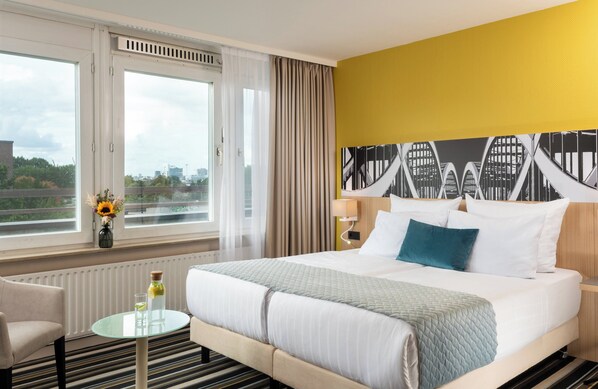 Habitación de hotel con una cama grande, una mesa de vidrio con una botella y un vaso, una silla, vistas de la ciudad a través de ventanas amplias y un cuadro de un paisaje urbano en la pared.